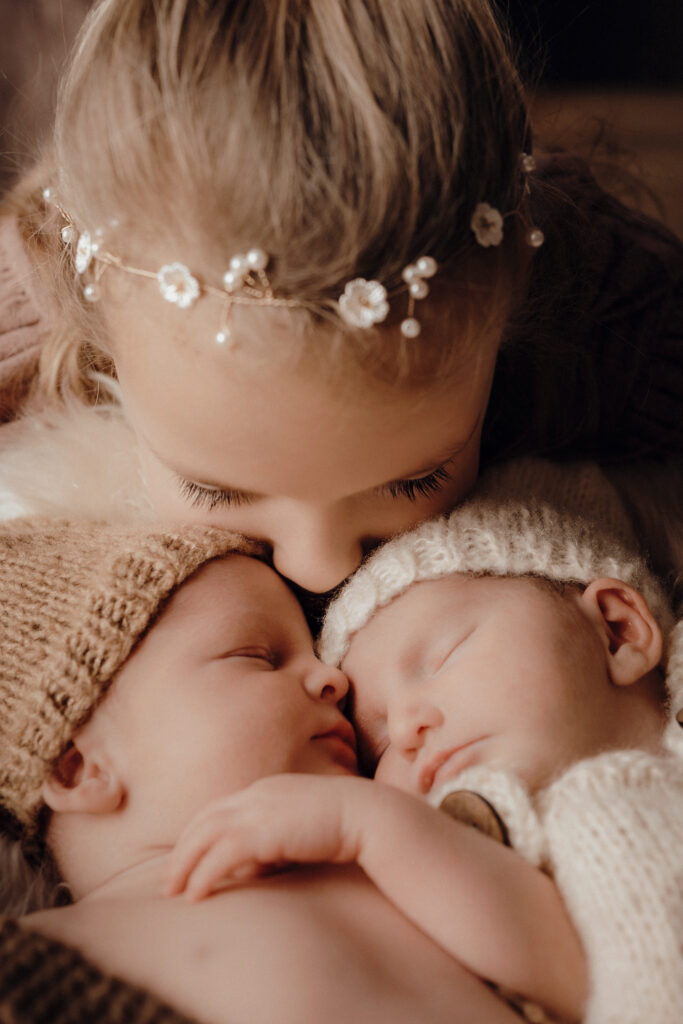 Große Schwester küsst die Zwillinge bei der liebevollen Neugeborenen-Fotografie bei einer Homesession. 