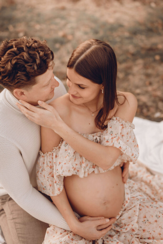 Schwangere lächelt den Vater liebevoll an und sitzt in der Natur beim Babybauchshooting